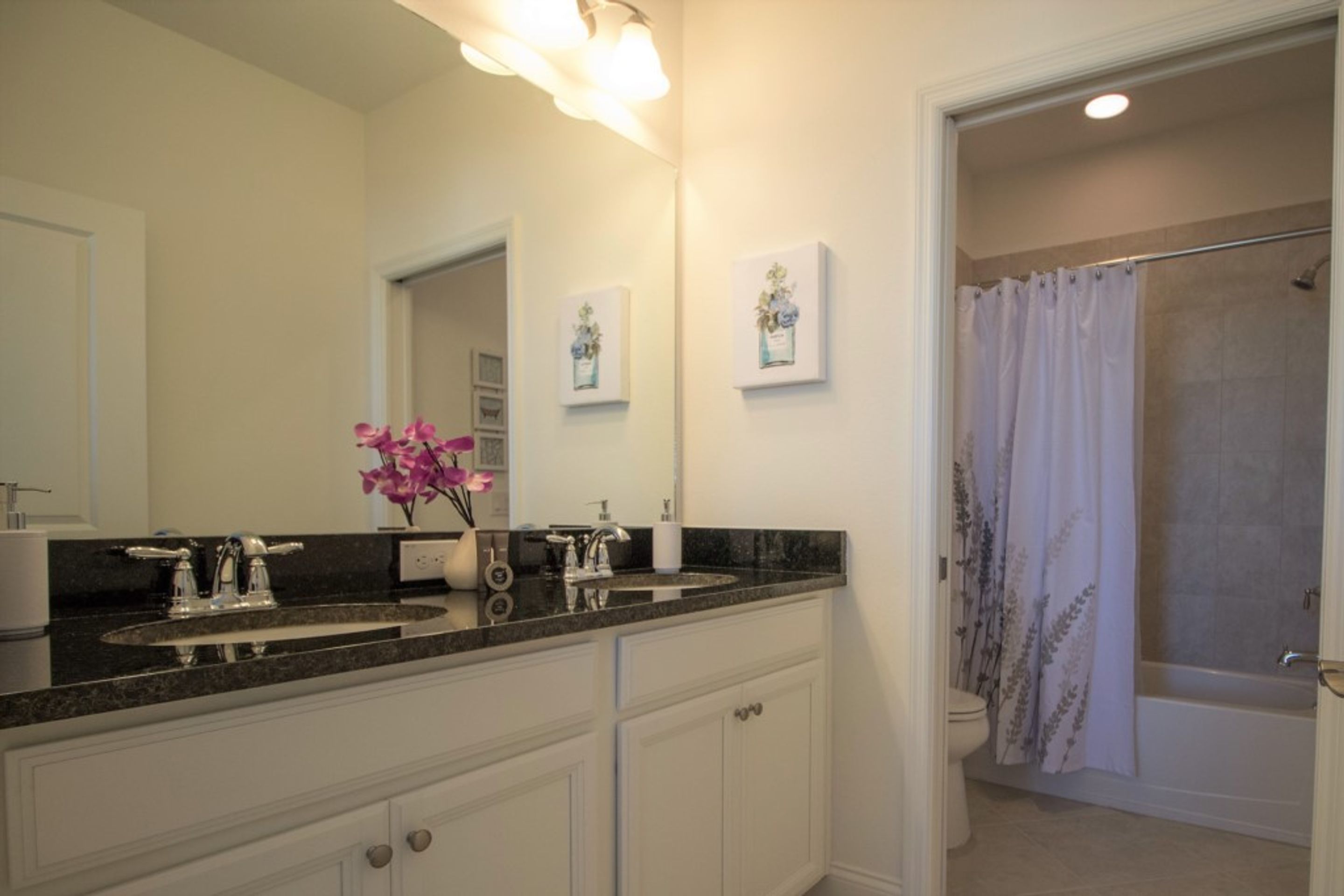 A bathroom with a double sink, a mirror, a shower with a floral curtain, and framed artwork on the wall.