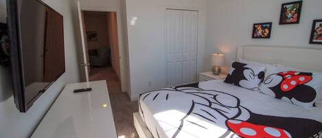 A bedroom with a bed featuring a Mickey Mouse design, a TV mounted on the wall, a bedside table with a lamp, and a door leading to another room.