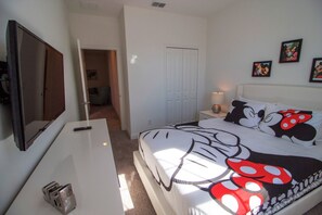 Ein Schlafzimmer mit einem Bett mit Mickey-Mouse-Motiv, einem an der Wand befestigten Fernseher, einem Nachttisch mit Lampe und einer Tür zu einem weiteren Raum.