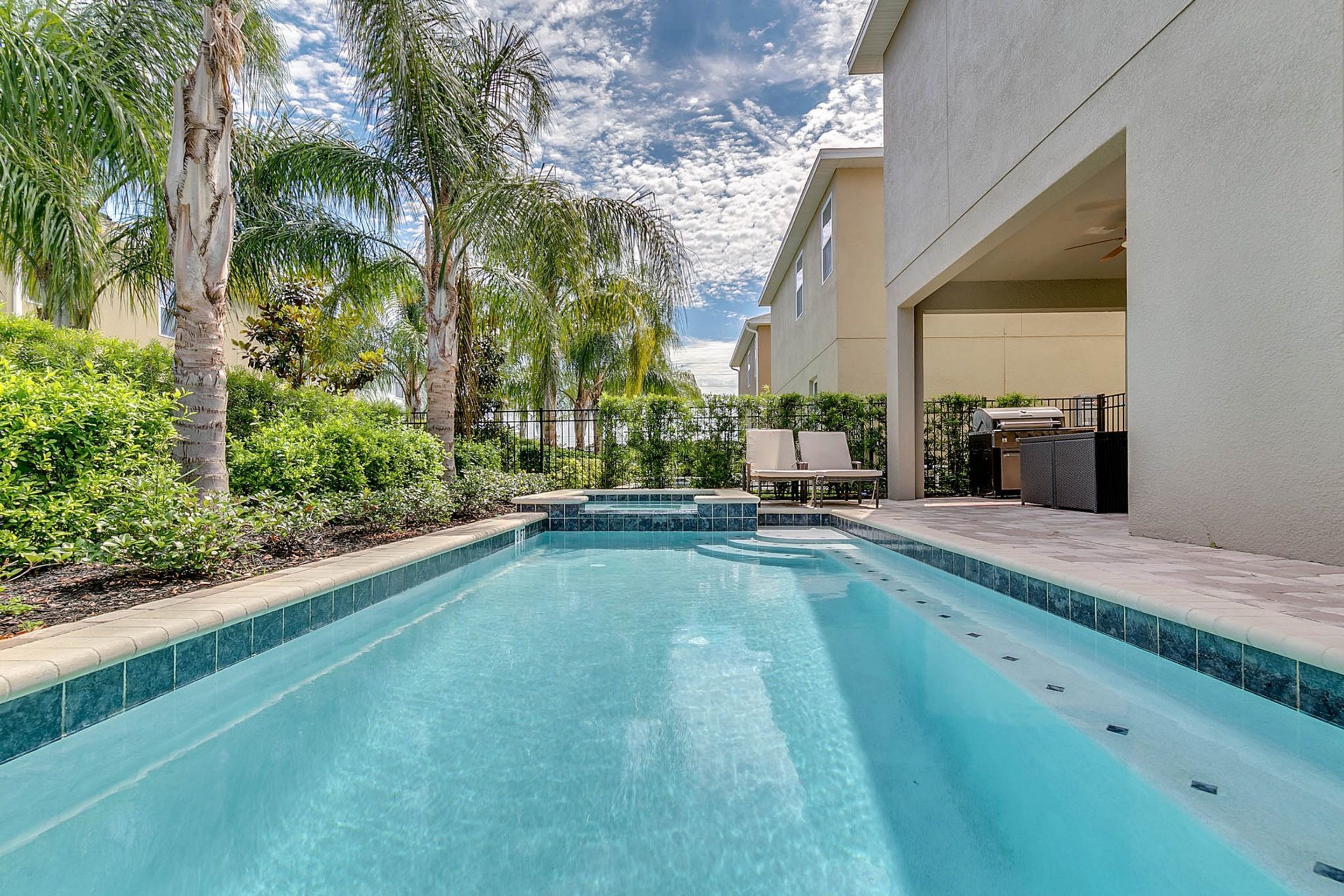 A swimming pool with a tiled edge, surrounded by palm trees and a building with a covered patio area.