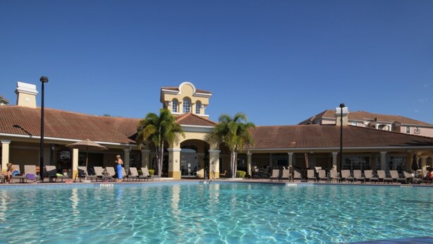 A swimming pool with lounge chairs and umbrellas, surrounded by a building with a tiled roof and palm trees. - Vista Cay 4840 - 108 (Orlando)