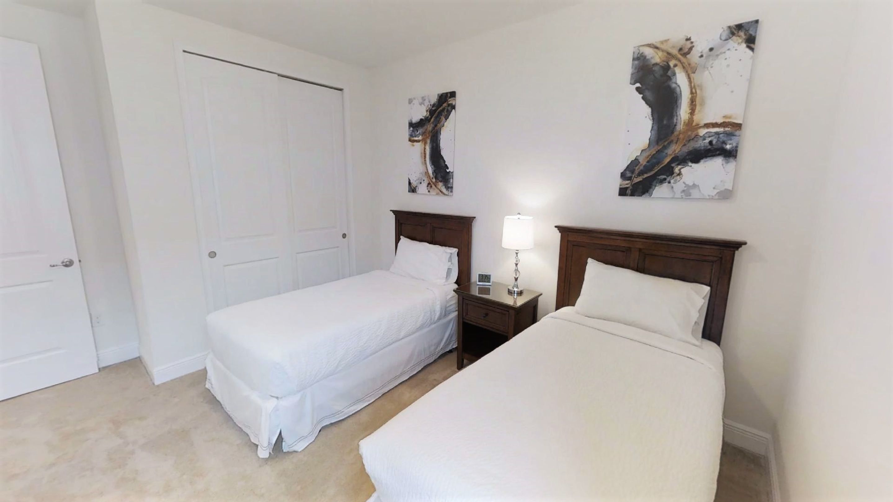 A hotel room with two single beds, a nightstand with a lamp, and abstract art on the walls.