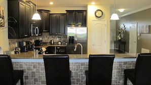 A modern kitchen with dark cabinets, a stainless steel refrigerator, a coffee maker, and a bar area with three chairs. - Champions Gate 1400 (Davenport)