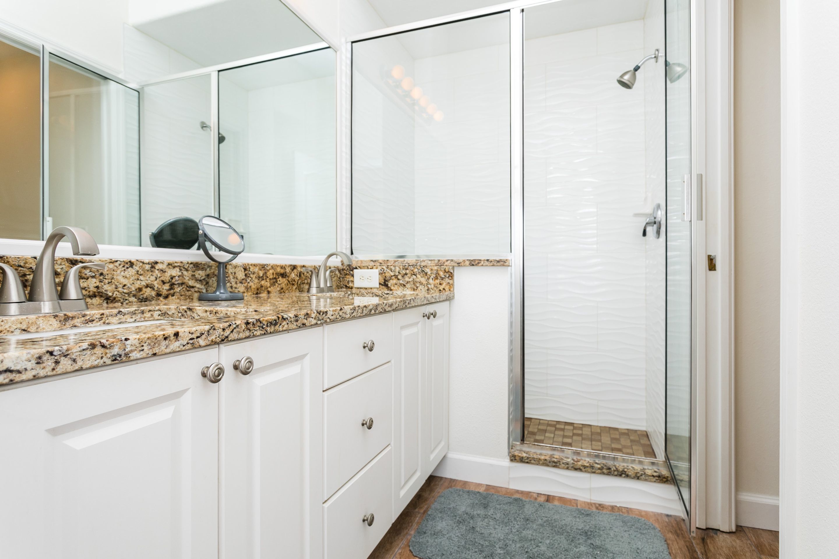 A modern bathroom with a granite countertop, white cabinets, a large mirror, a shower with a glass door, and a small rug on the floor.