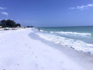 A beach with white sand, clear blue water, and a few umbrellas.