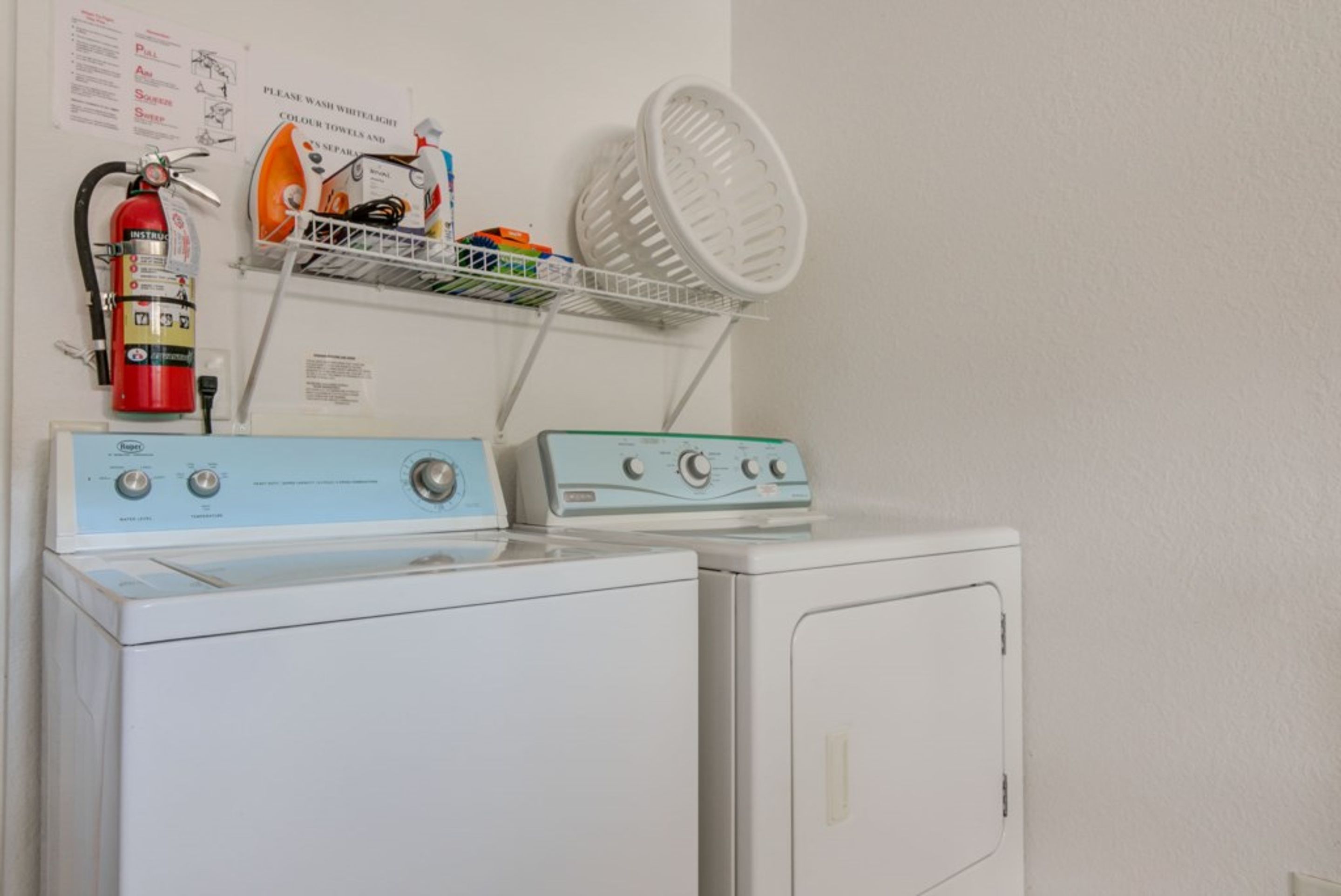 Un espace buanderie équipé de deux machines à laver, d’un extincteur et d’une étagère contenant des produits de nettoyage.