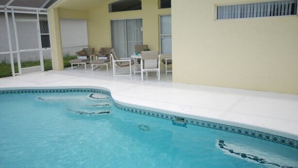 Une piscine avec un bord carrelé, un patio couvert avec des chaises, et un bâtiment à l’extérieur jaune.