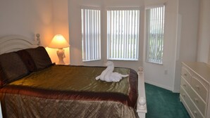 Une chambre à coucher comprenant un lit, une table de chevet, une lampe, une commode et deux fenêtres équipées de stores.