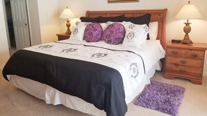 Ein ordentlich bezogenes Bett mit grau-weiß gestreifter Tagesdecke, zwei Nachttische mit Lampen, ein violettes Teppichläufer und ein gerahmtes Bild an der Wand.