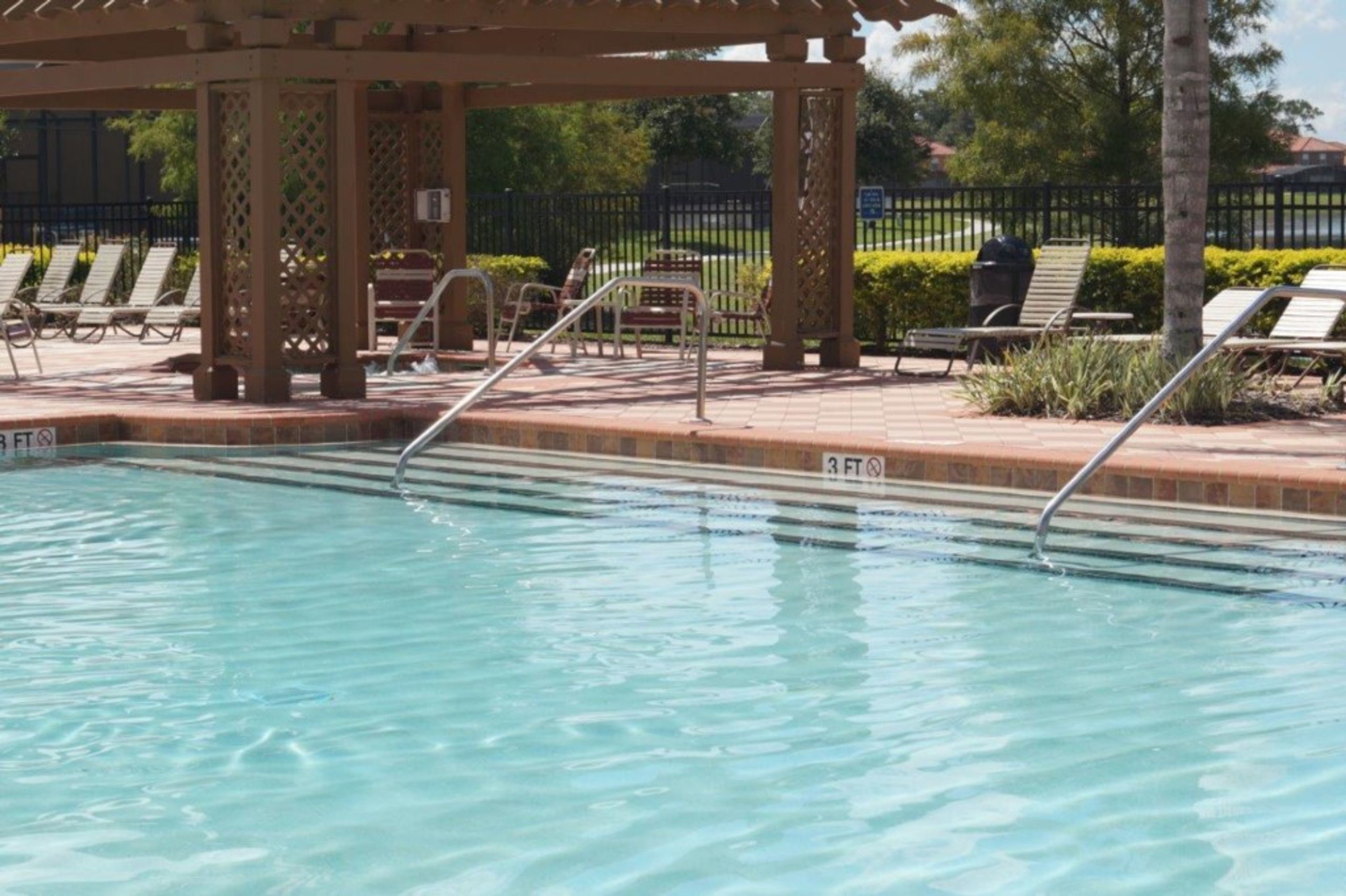 A swimming pool with a wooden pergola, lounge chairs, and a fenced area with greenery.