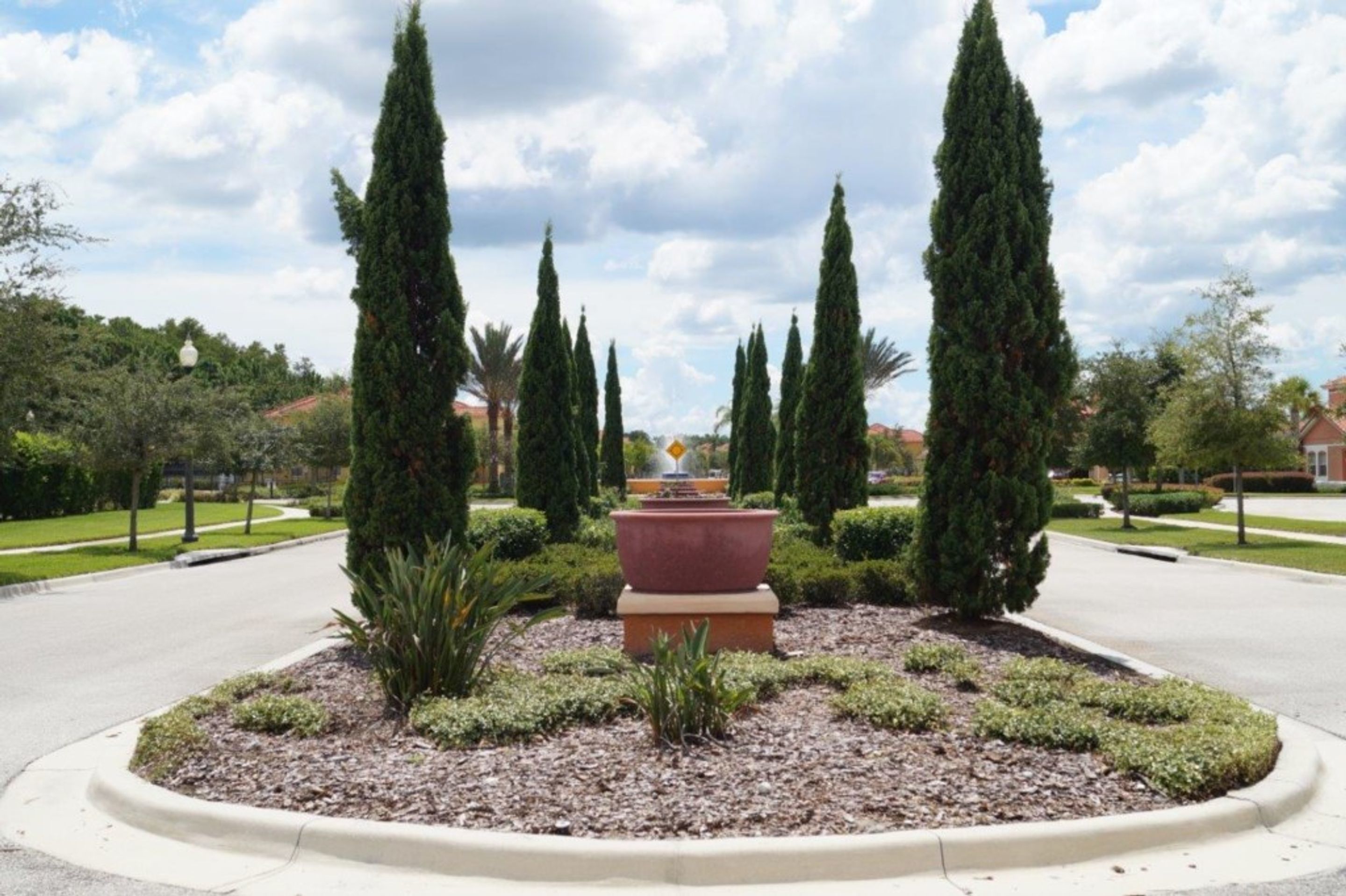 A landscaped garden with a central fountain, surrounded by neatly trimmed hedges and tall cypress trees.