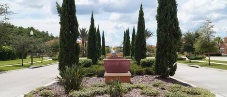 A landscaped garden with a central fountain, surrounded by neatly trimmed hedges and tall cypress trees.