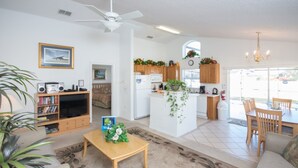 A spacious living room with a sofa, a coffee table, and a TV. There is a kitchen area with wooden cabinets and a dining area with a table and chairs.