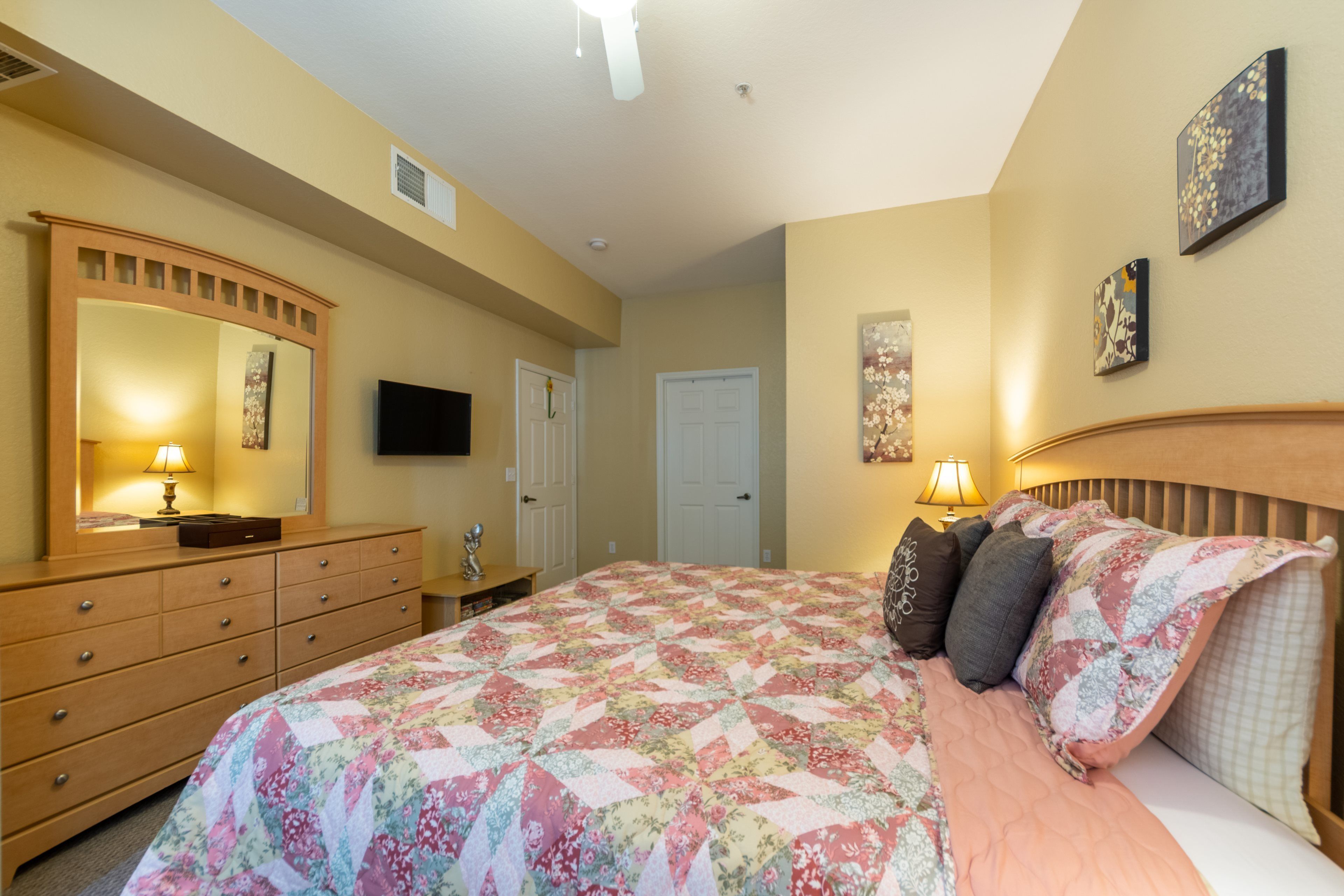 A bedroom with a large bed, a dresser, a mirror, a flat-screen TV, and two doors.