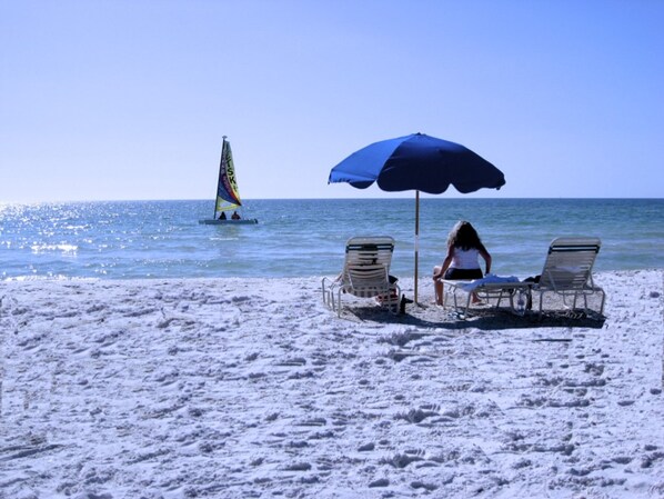 A beach with two lounge chairs under a blue umbrella, a sailboat in the water, and a clear sky.