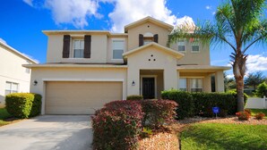 A two-story house with a beige facade, a garage, and a well-maintained front yard with a palm tree and flowering bushes. - Legacy Park 120 (Davenport)