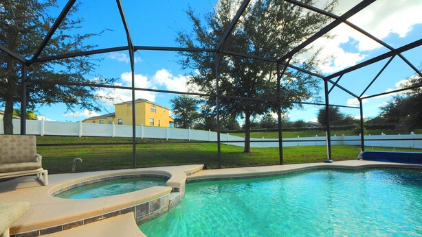 A swimming pool with a hot tub, surrounded by a covered area with a view of a yellow house and a white fence in the background. - Legacy Park 120 (Davenport)