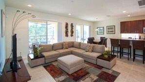 Una sala moderna con un sofá modular, una mesa de centro y un televisor. Se aprecia un comedor con sillas y una mesa. La cocina es visible, con armarios oscuros y una barra.