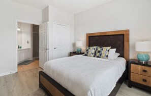A bedroom with a large bed, wooden headboard, two bedside tables with lamps, and a door leading to a bathroom. - Vista Cay 4862 - 202 (Orlando)