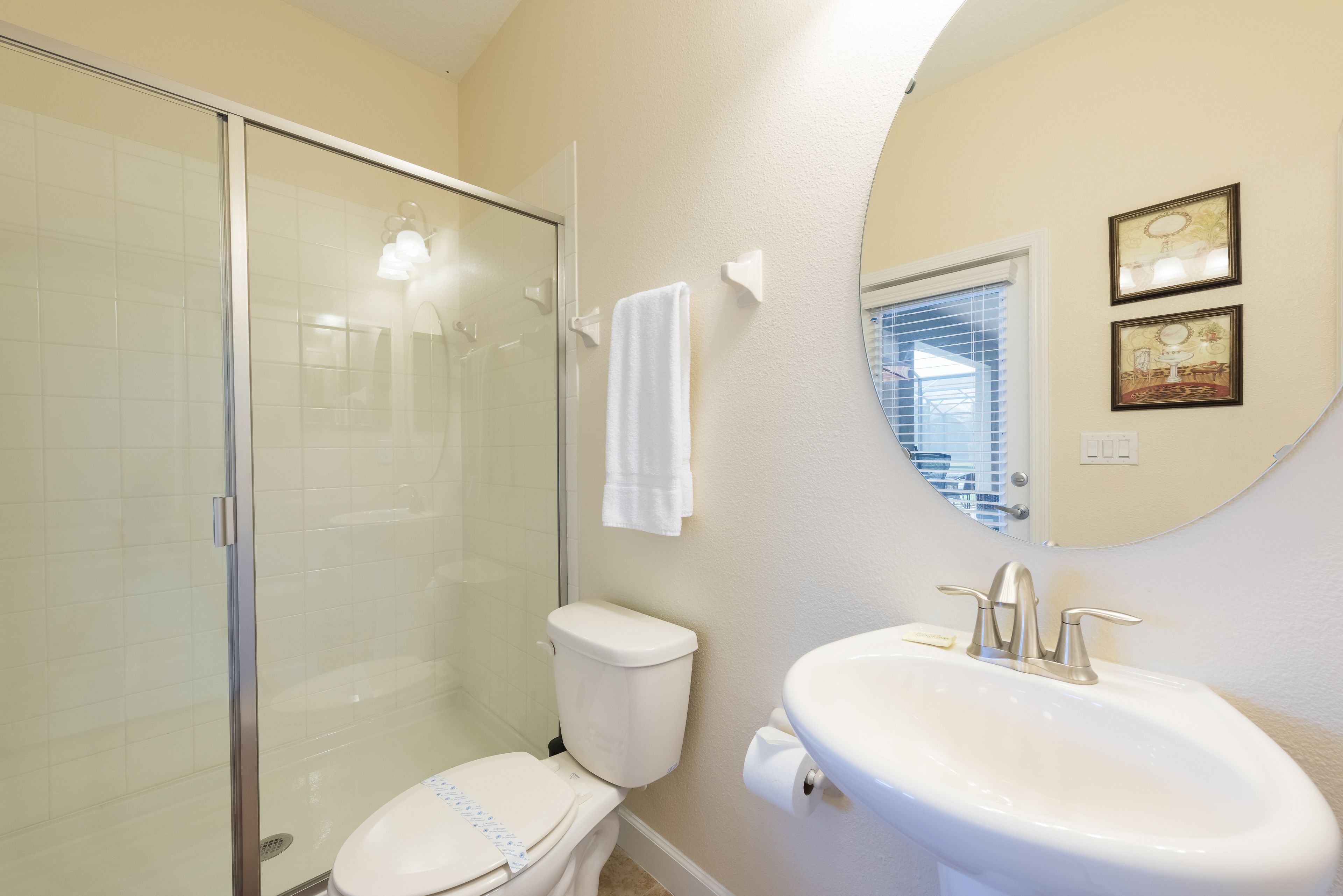 A bathroom with a glass shower enclosure, a toilet, a sink with a mirror, and two framed pictures on the wall.