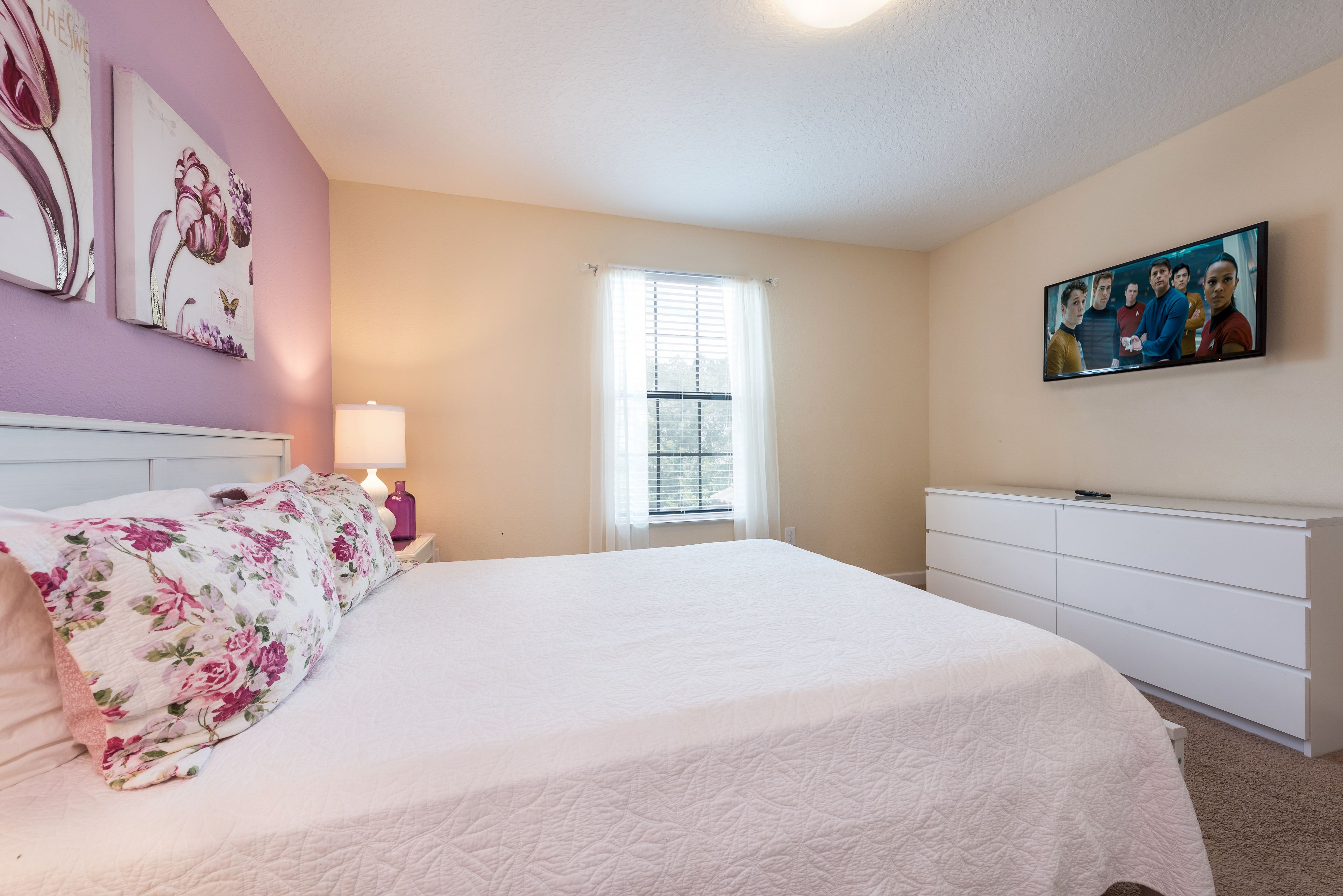 A bedroom with a bed, a nightstand, a dresser, a window with blinds, and two wall-mounted paintings.