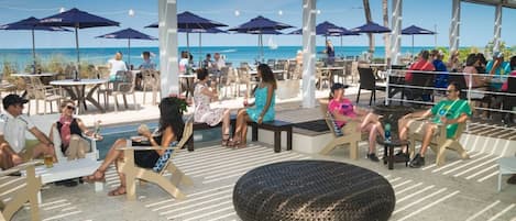 An outdoor seating area with tables and chairs, umbrellas, and a view of the beach.