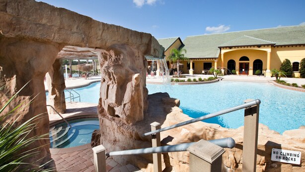 A pool area with a rock formation, a sign prohibiting climbing on rocks, and a building with a tiled roof in the background. - Paradise Palms 8964 (Kissimmee)
