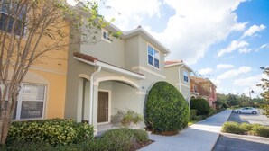 A two-story residential building with a light-colored facade, a covered front porch, and a well-maintained front yard with a trimmed bush and a tree. - Paradise Palms 8964 (Kissimmee)
