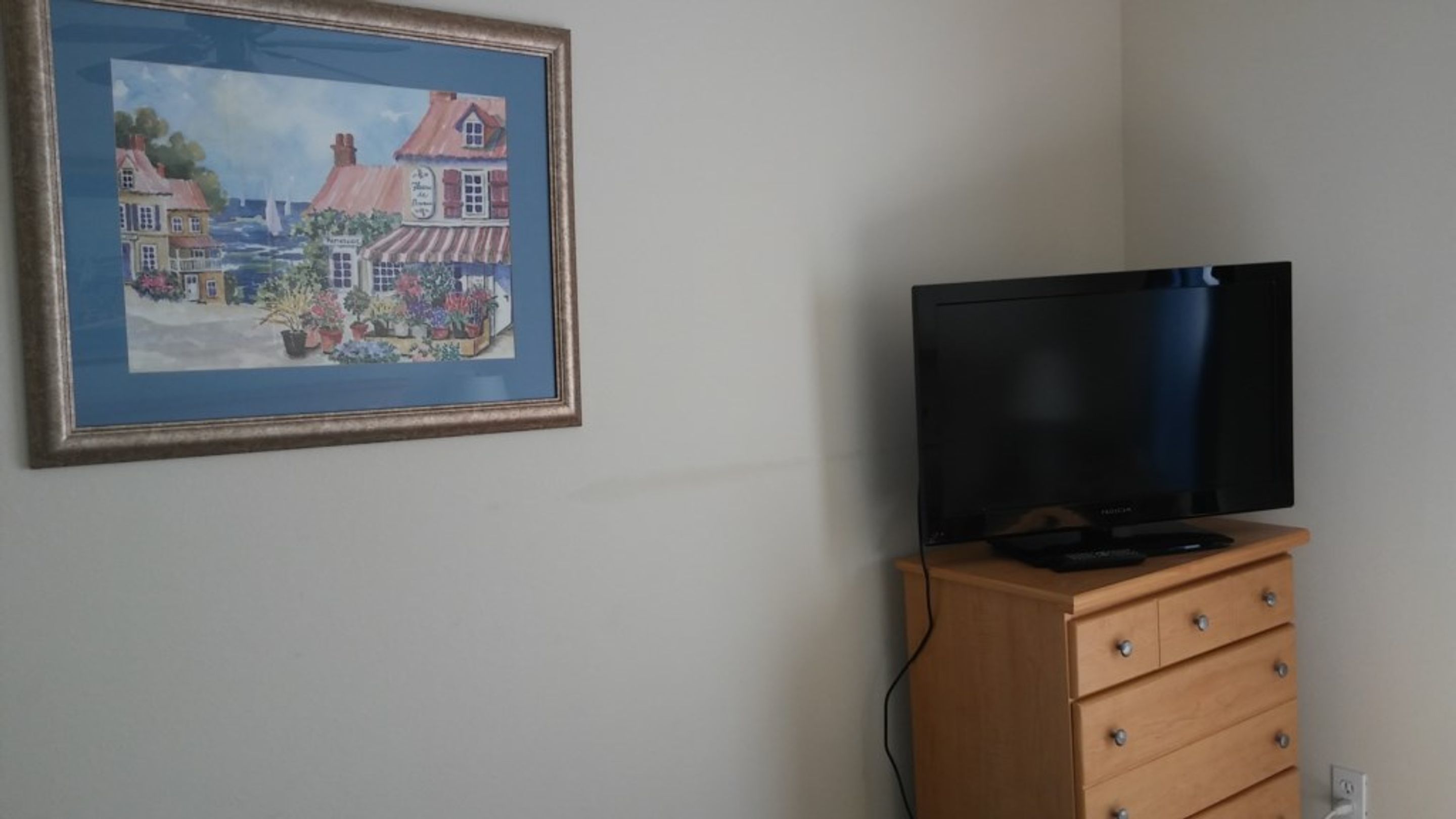 A room with a framed painting of a town scene, a wooden dresser with a flat-screen TV, and a power outlet on the wall.