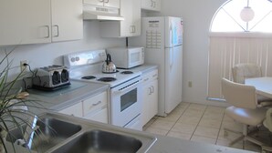 A kitchen with white cabinets, a stove, a microwave, and a refrigerator. There is a window with blinds and a plant in the corner. - Indian Wells 5356 (Kissimmee)