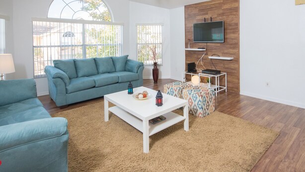 Un salon avec un canapé bleu, une table basse blanche et un pouf à motifs. Un téléviseur est fixé au mur en bois, il y a un ventilateur de plafond et de grandes fenêtres donnant sur des arbres.