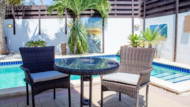 A poolside area with wicker chairs and a glass table, surrounded by palm trees and a blue-tiled pool. - Noord 2 (Noord)