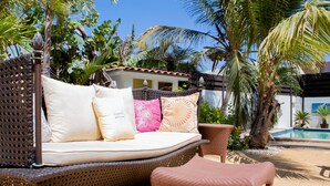 A wicker sofa with cushions and a matching ottoman in a tropical outdoor setting with a pool. - Noord 2 (Noord)
