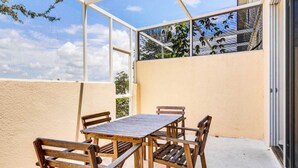 Een overdekt terras met een houten tafel en stoelen, grote ramen en uitzicht op de lucht en bomen.