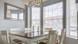 Een eetruimte met een houten tafel, witte stoelen, grote ramen met zonwering en een spiegel aan de muur.
