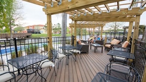 Une terrasse extérieure avec des tables et des chaises, une pergola en bois et une vue sur une étendue d’eau et des bâtiments en arrière-plan.