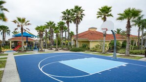 Un terrain de basket en plein air, doté d’une surface bleue, entouré de palmiers et comprenant une aire de jeux avec un toboggan rouge.