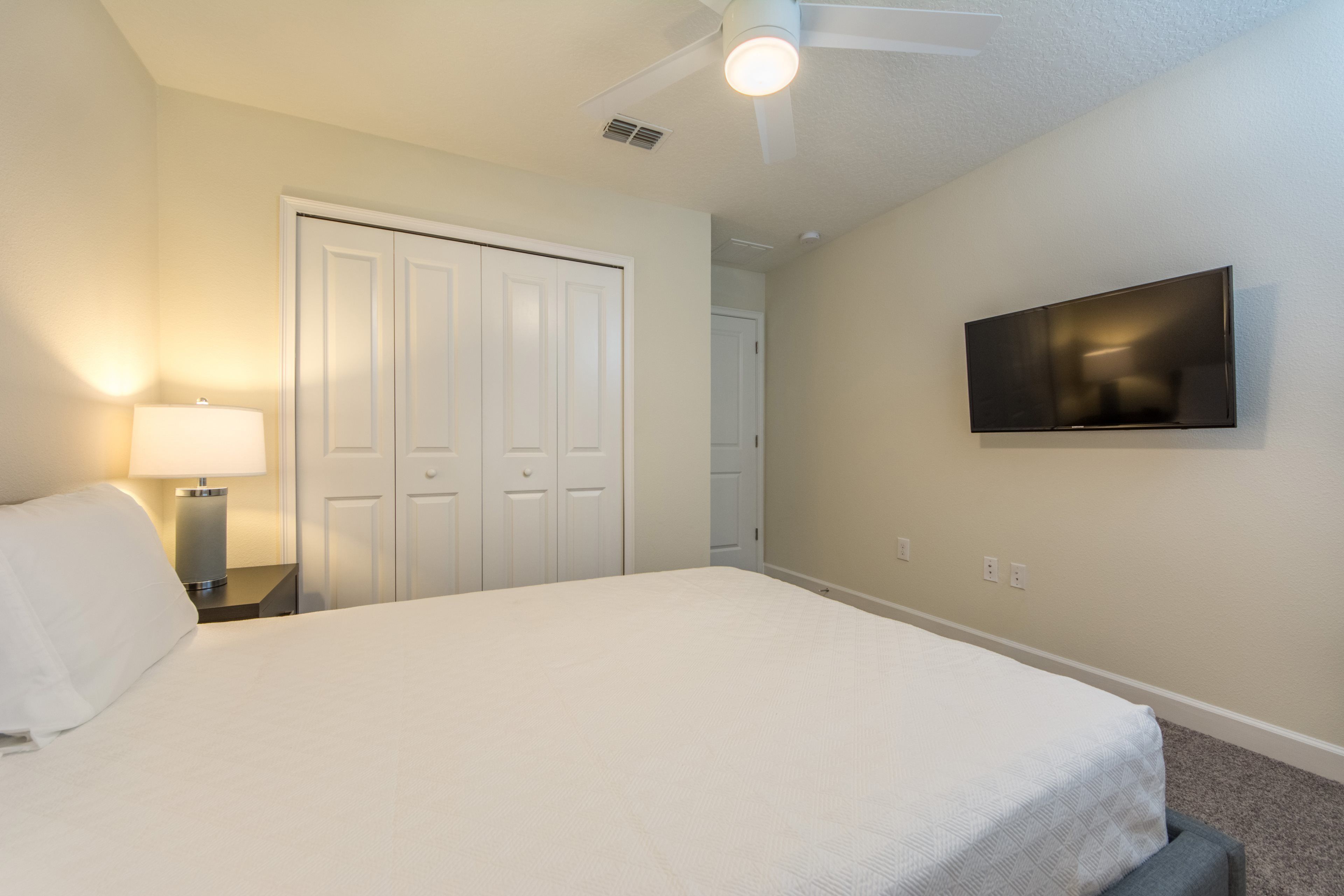 Un dormitorio con cama, televisión montada en la pared, ventilador de techo y puerta con espejo.