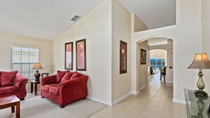 A spacious living room with a red sofa set, a coffee table, and a lamp. There are framed pictures on the walls and a view into the dining area through an archway. - Rolling Hills 7979 (Kissimmee)