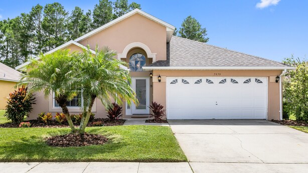 A single-story house with a beige exterior, a white garage door, and a well-maintained lawn with palm trees. - Rolling Hills 7979 (Kissimmee)