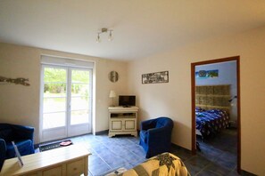 Ein Zimmer mit einer großen Glasschiebetür, die auf einen Balkon führt, einem kleinen weißen Schrank mit einem Fernseher, einem blauen Sessel und einem Bett mit einer blau-weißen Bettdecke.
