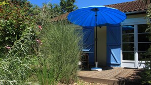 Een houten terras leidt naar een huis met een blauwe parasol, omringd door groen en een helderblauwe lucht.