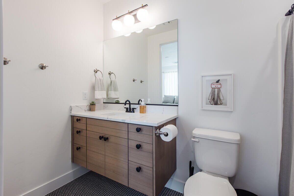 Une salle de bain avec un lavabo blanc, un meuble en bois, un tapis de sol noir, des toilettes blanches et un miroir avec éclairage intégré.