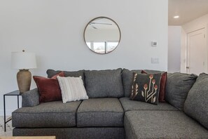 Ein graues Ecksofa mit vielen Kissen, ein runder Spiegel an der Wand, ein kleiner Tisch mit einer Lampe und eine Tür im Hintergrund.