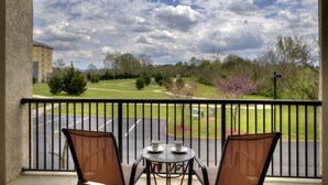 A balcony with two chairs and a table, overlooking a parking lot and a grassy area with trees and a building in the background. - Updated Clean Spacious 3BR/2BA Condo right off the parkway! Pools/WiFi/Grills (Pigeon Forge)