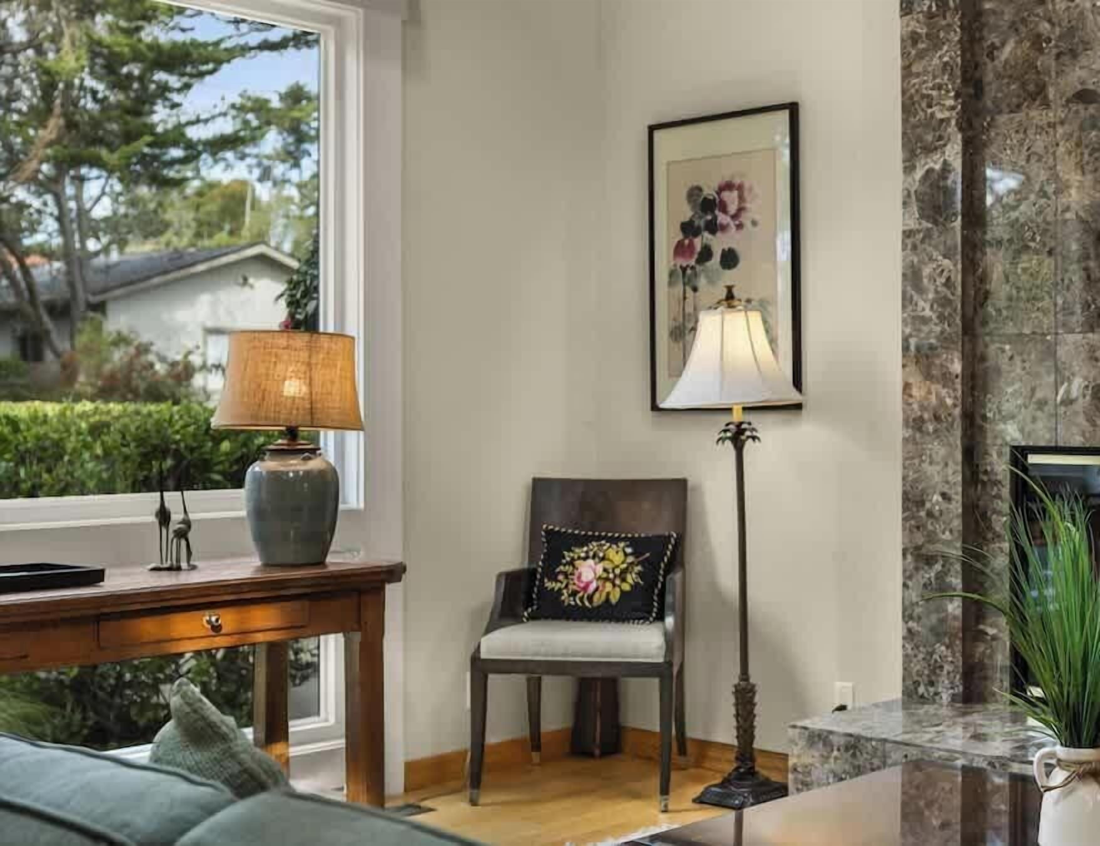A cozy living room with a wooden desk, a chair with a floral cushion, a floor lamp, and a framed artwork on the wall.