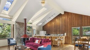 Ein geräumiger Wohnbereich mit einem roten Sofa, einem Couchtisch und einem Kamin. Es gibt eine Küchenzeile mit Holzausstattung und einen Esstisch. Große Fenster sorgen für natürliches Licht und Ausblicke ins Freie.