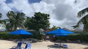 En strand med liggestole og blå parasoller, et skilt for Buccaneer Beach Club, og en privat parkeringsplads.