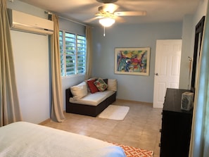 A bedroom with a bed, a sofa bed, a ceiling fan, a window with curtains, a door, and a painting on the wall.