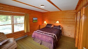Ein Schlafzimmer in einer Blockhütte mit einem Bett, zwei Nachttischlampen, einem Fenster mit Vorhängen und Blick ins Freie.